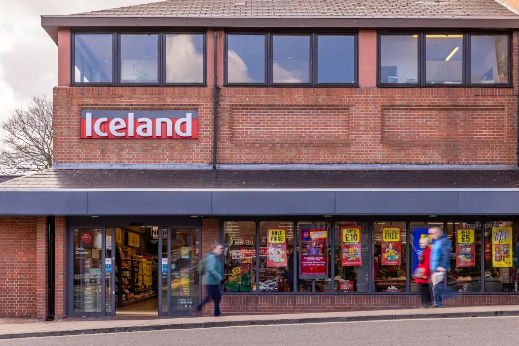 The front of an Iceland grocery store, featuring red signage and shoppers passing by, with colorful sale signs visible in the windows.