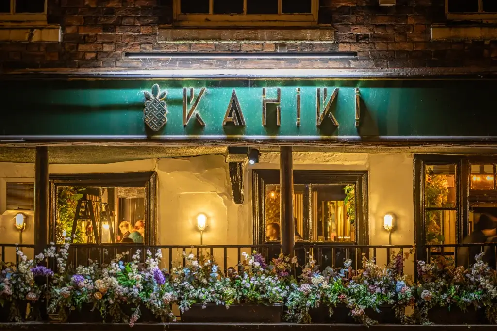 A warmly lit restaurant facade with the sign "Kahiki" and a vibrant floral display. Inside, people are seen interacting.