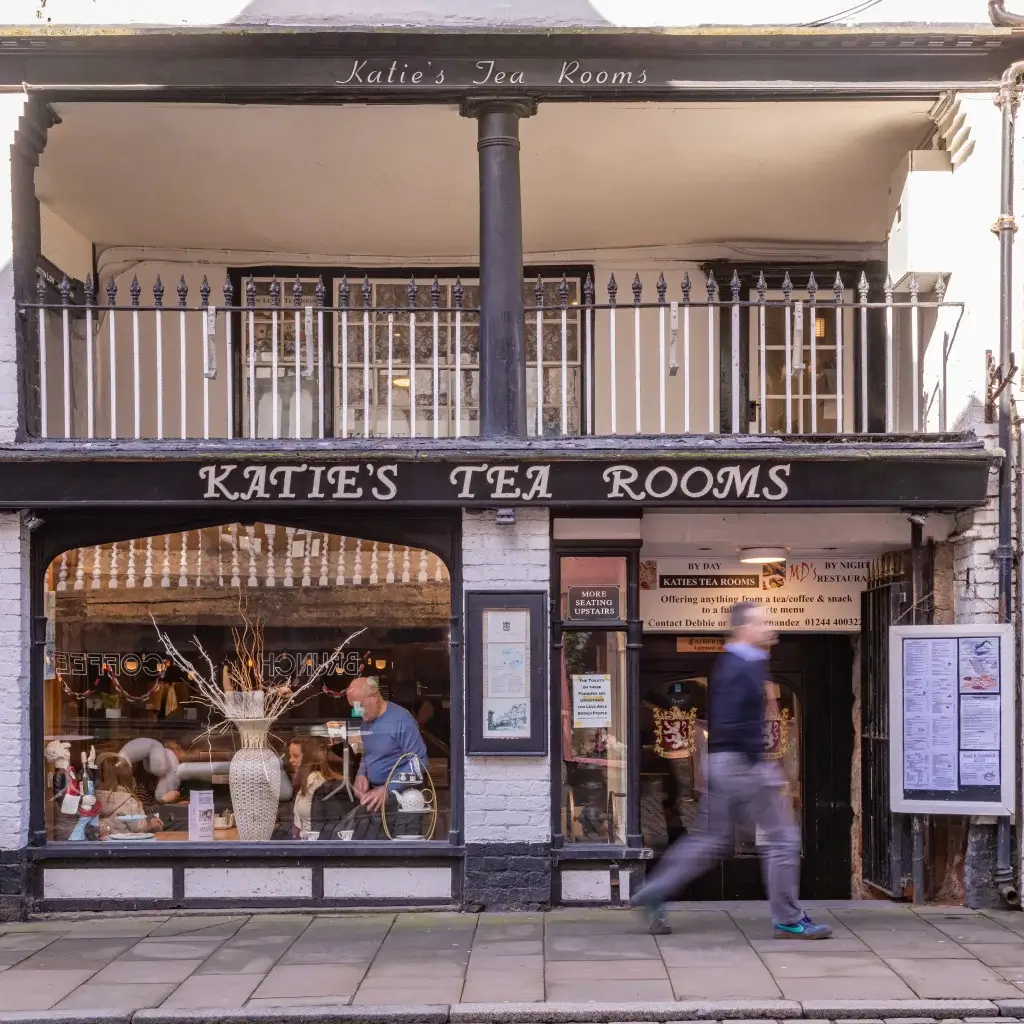 A pedestrian walks past Katie's Tea Rooms, featuring a warm interior with tables and decorative elements in the windows.