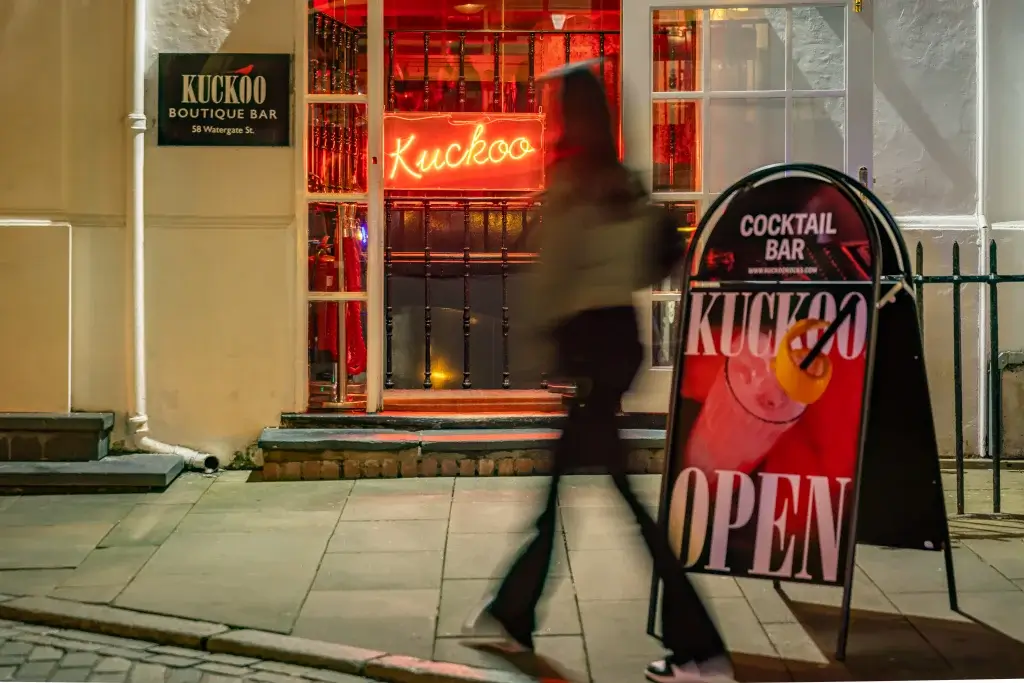 A blurred figure walks past the illuminated Kuckoo Boutique Bar, showcasing a neon sign and an open cocktail bar display outside.