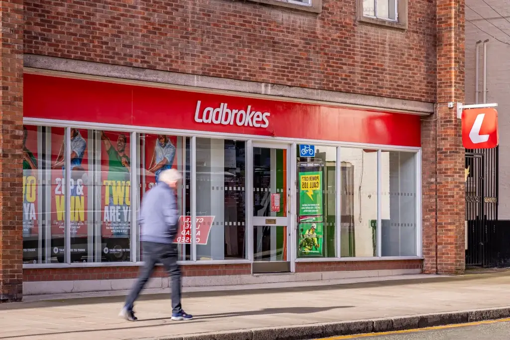 A person walks past a Ladbrokes betting shop, featuring bright signage and promotional posters in the windows.