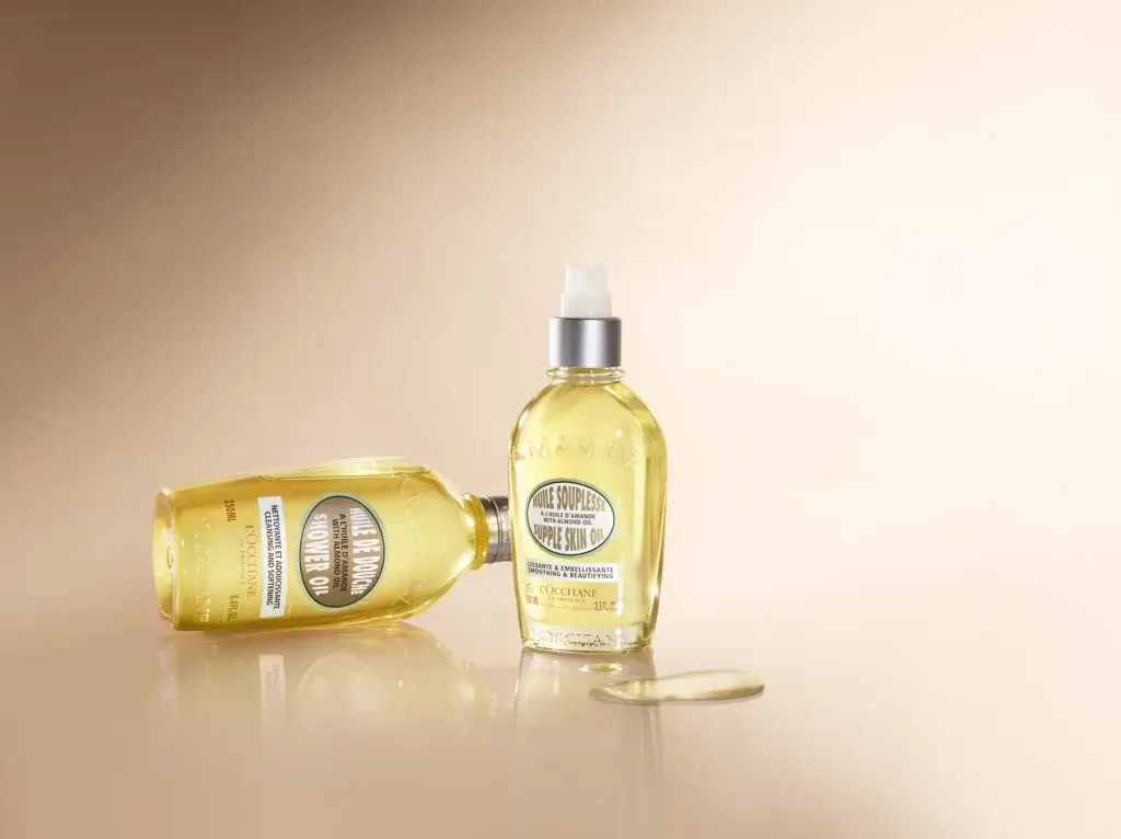 Two bottles of L'Occitane skin oil, one upright and one tipped, with a glossy surface and warm beige background.
