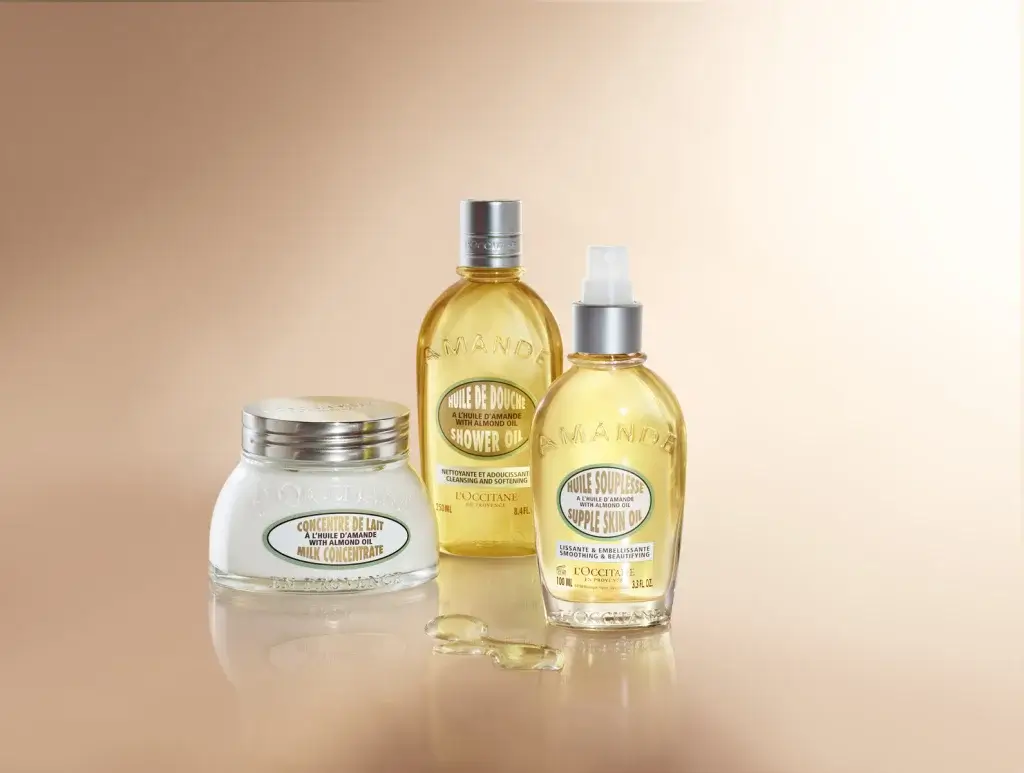 Three L'Occitane almond products: Shower Oil, Supple Skin Oil, and Milk Concentrate, arranged elegantly against a soft gradient background.