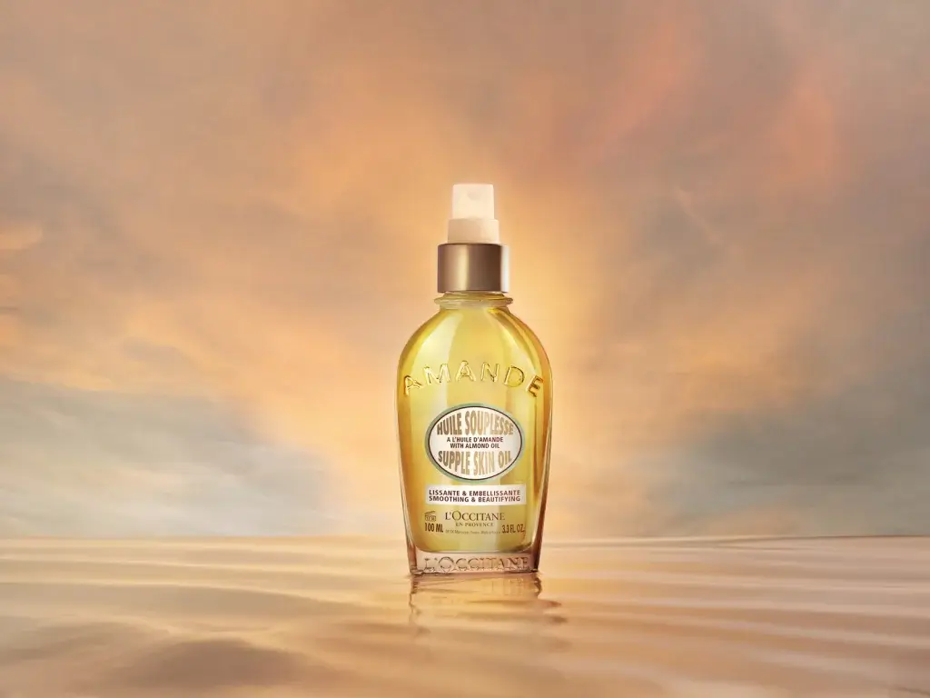A bottle of L'Occitane Amande Almond Oil against a soft, colorful sunset backdrop, reflecting on calm water.