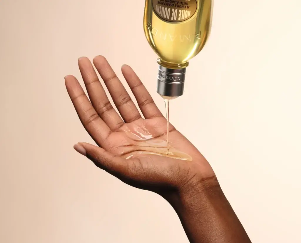 A hand extended upward, catching drops of golden oil from a bottle against a soft, neutral background.