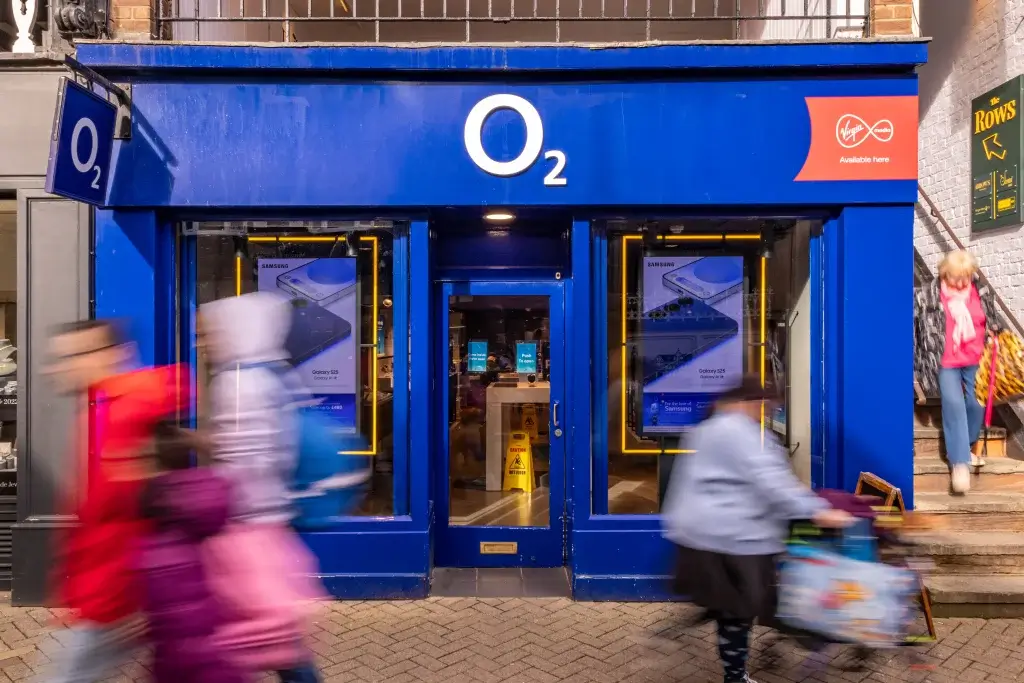 A busy O2 store entrance with vibrant blue walls, featuring Samsung advertisements, as pedestrians hurry past.