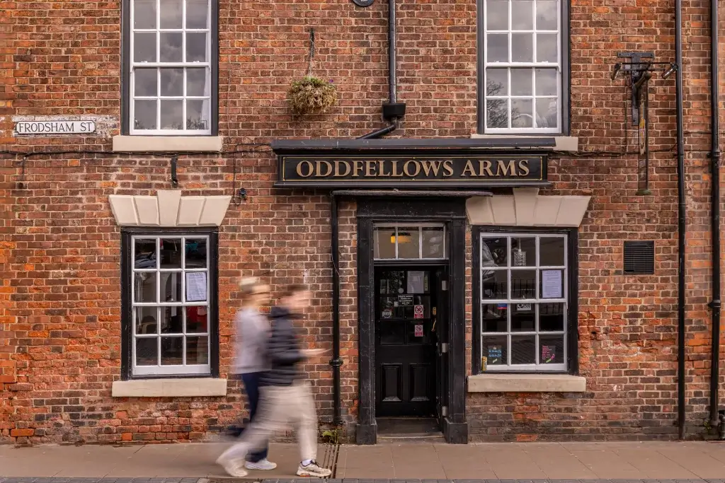 The brick facade of Oddfellows Arms pub, with a street sign for Frodsham St, and blurred figures walking by.