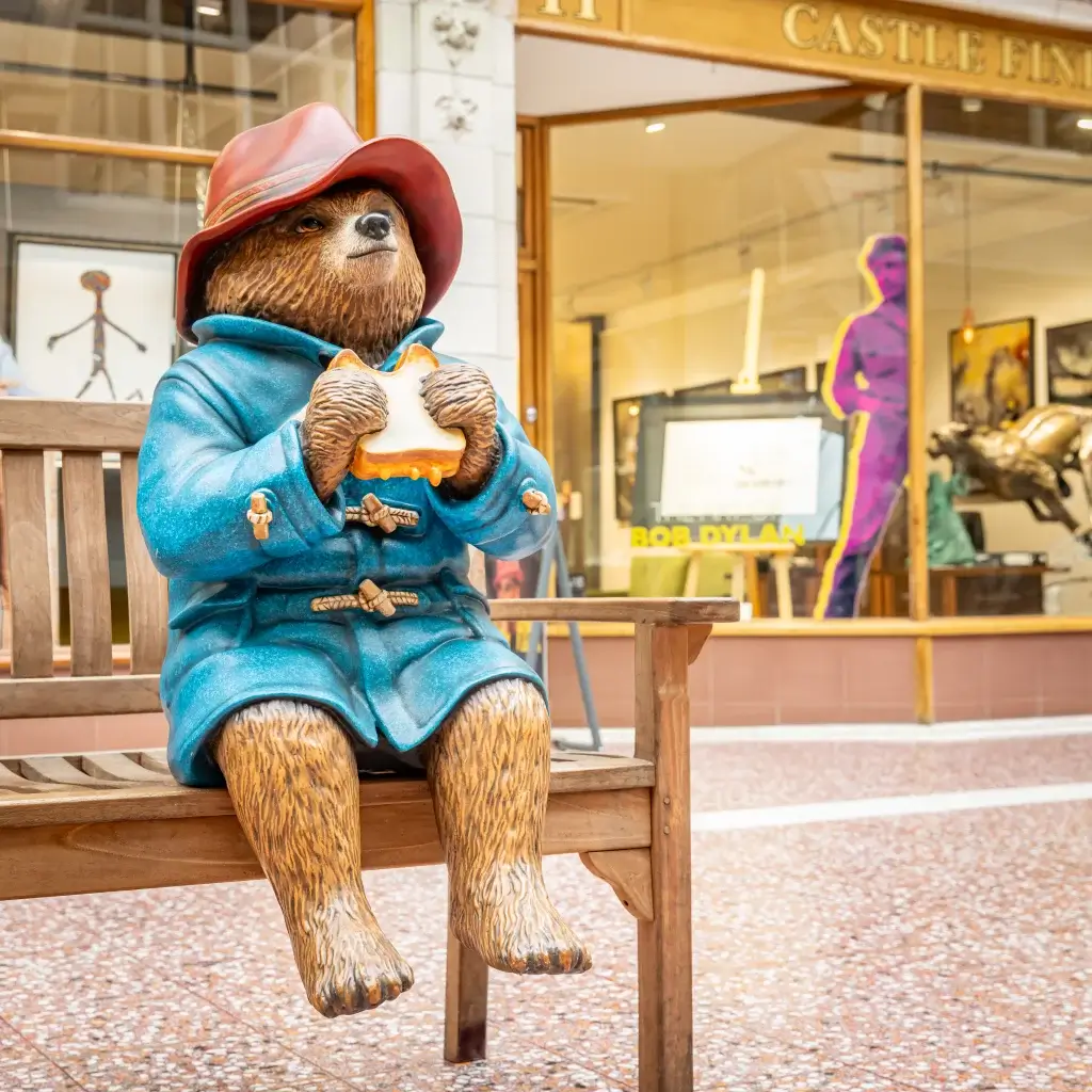 A charming statue of a bear wearing a red hat and blue coat, sitting on a wooden bench and holding a sandwich, surrounded by art displays.