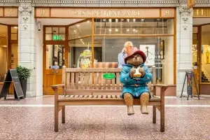 A charming statue of a bear wearing a red hat and blue coat, sitting on a wooden bench and holding a sandwich, surrounded by art displays.