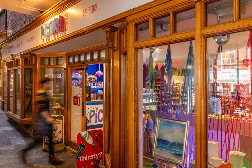 Wood-framed art school storefront with colourful interior, a displayed seascape painting and bunting, and a blurred pedestrian passing by.