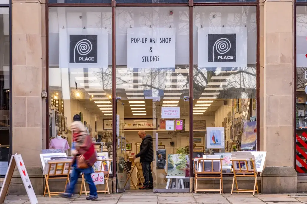 Pop-up art shop and studios shopfront with paintings on easels outside, a person browsing inside and a blurred passerby.