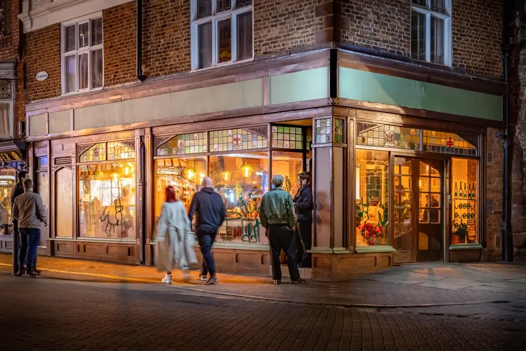 People walk past a warmly lit corner restaurant with ornate windows and inviting interior, set in a charming brick building at night.
