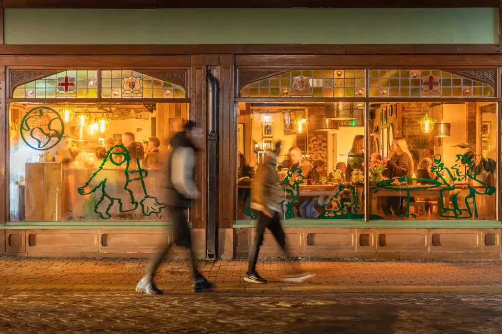Two pedestrians walk past a brightly lit restaurant window featuring colorful wall art and people enjoying their meals inside.