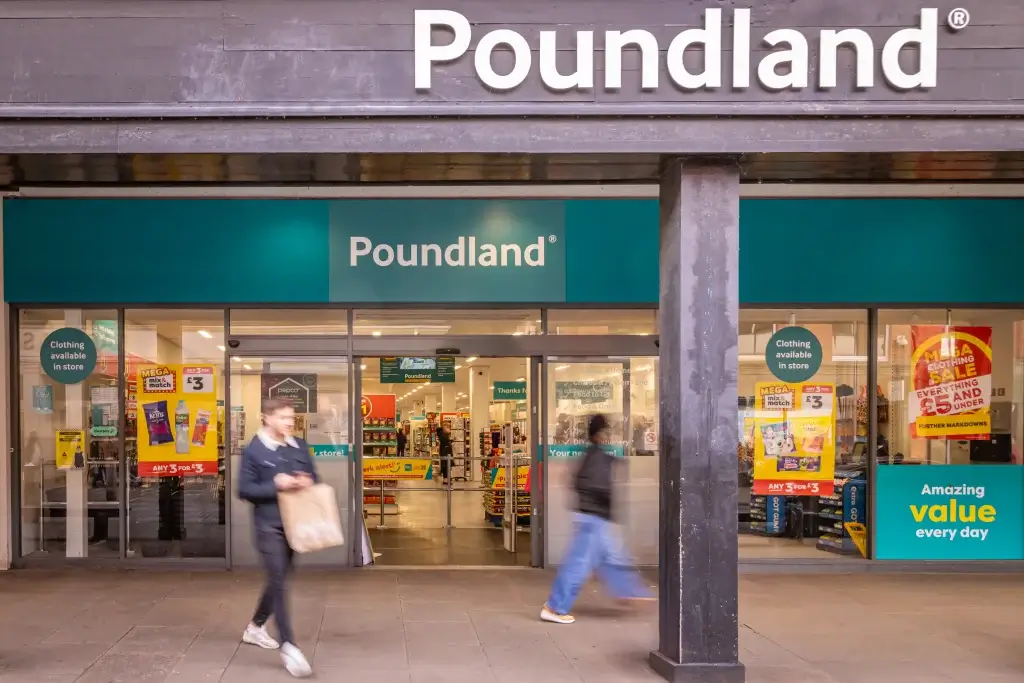 A storefront of Poundland features bright signage, promotional posters, and shoppers entering and exiting the store.
