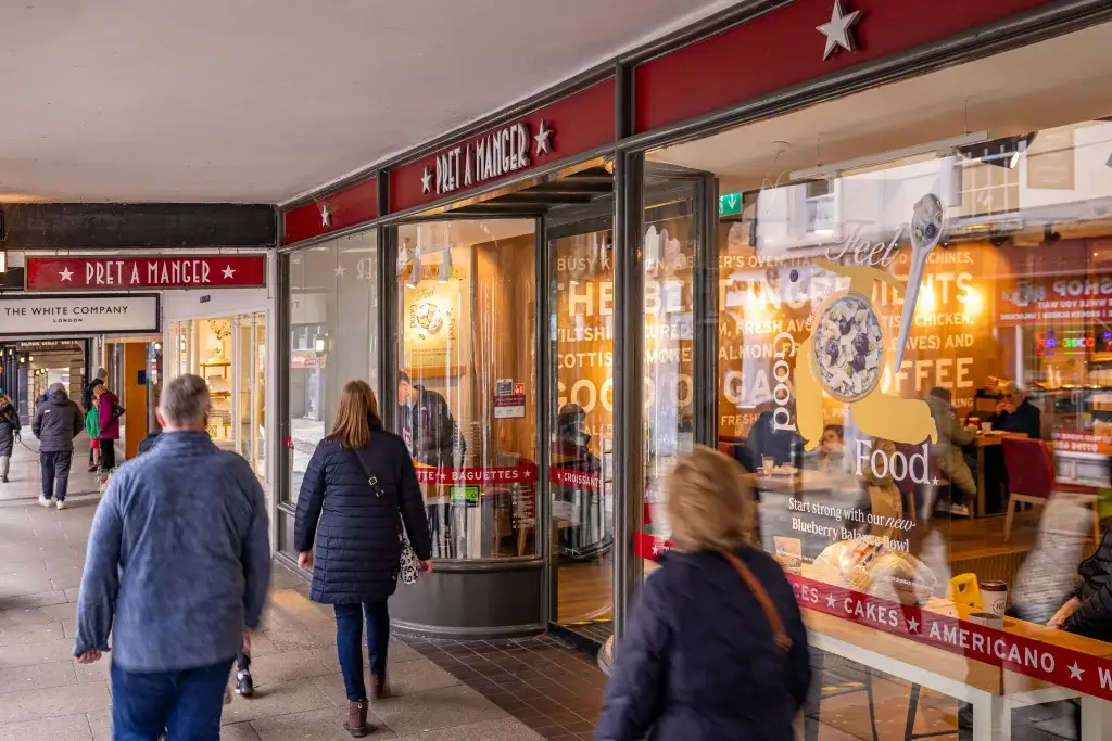 A Pret A Manger café storefront with people walking past, showcasing the wooden architecture and displays of sandwiches and drinks inside.