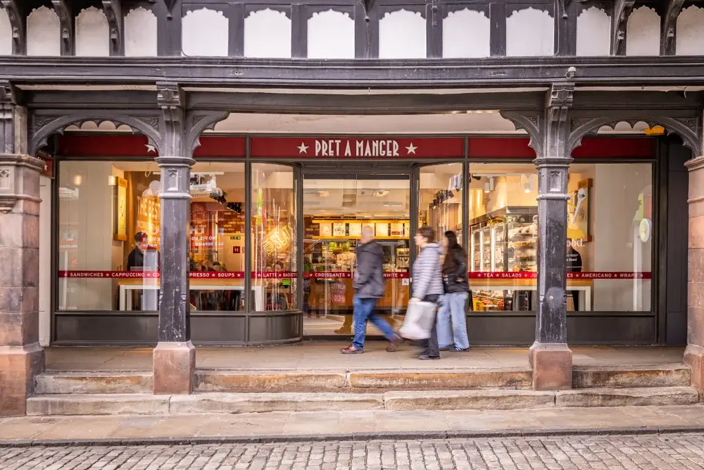 A Pret A Manger café storefront with people walking past, showcasing the wooden architecture and displays of sandwiches and drinks inside.