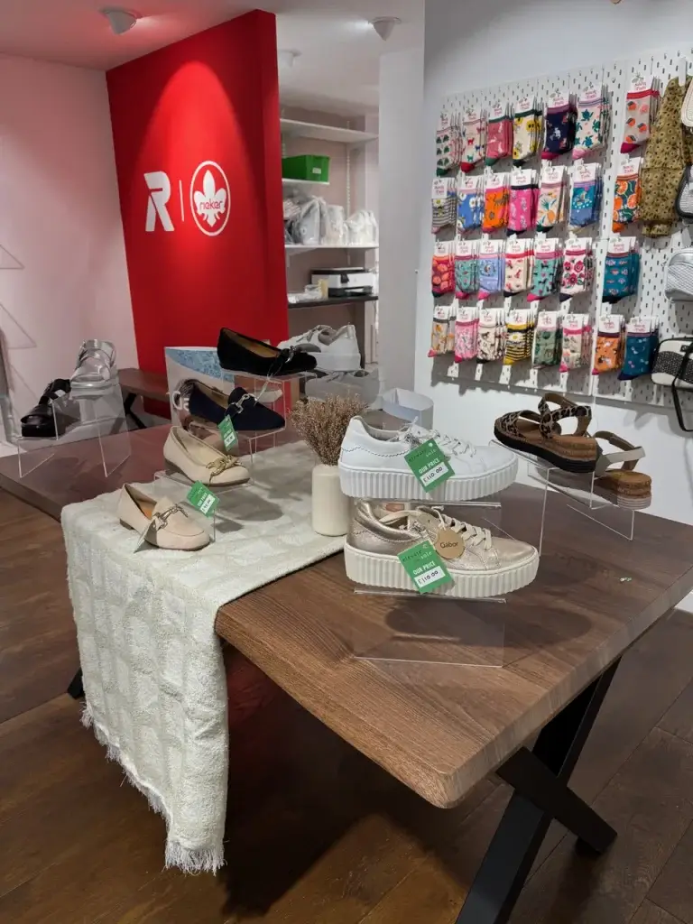 A display of stylish shoes on a wooden table, surrounded by colorful socks hanging on the wall, featuring the brand logo in the background.