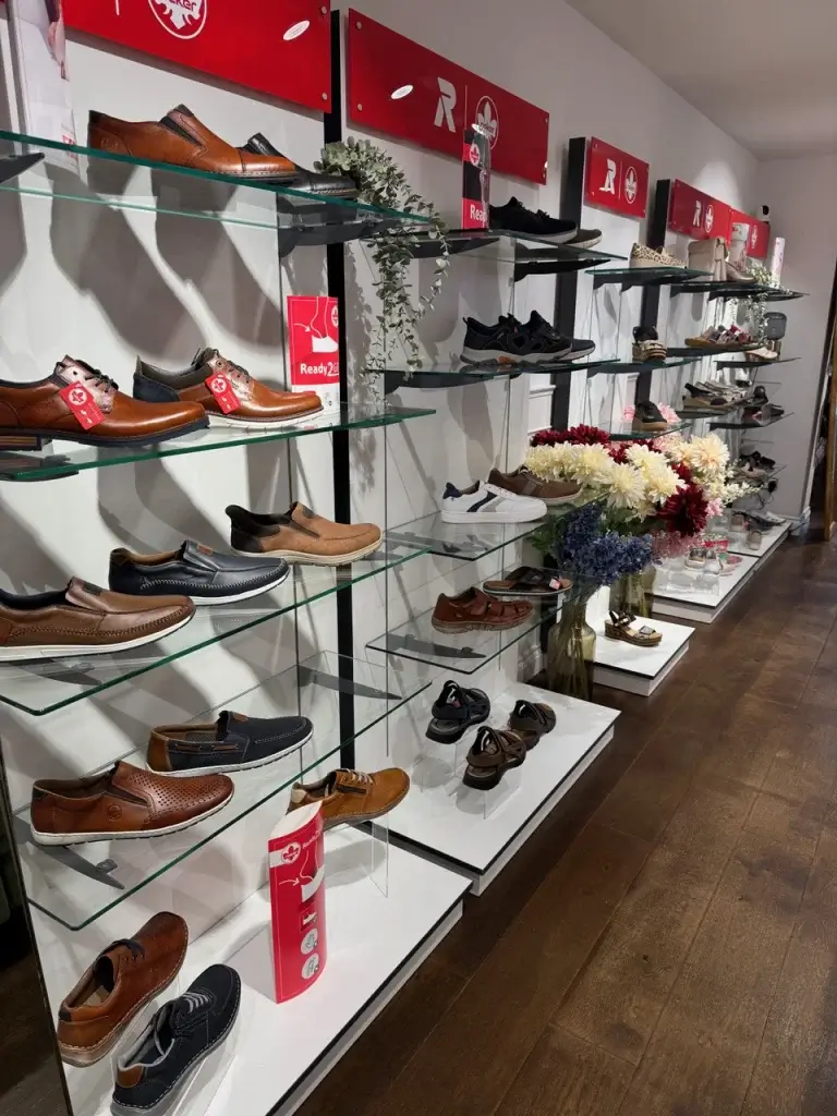 A variety of men's shoes displayed on glass shelves, featuring diverse styles and colors, with floral decor in the background.