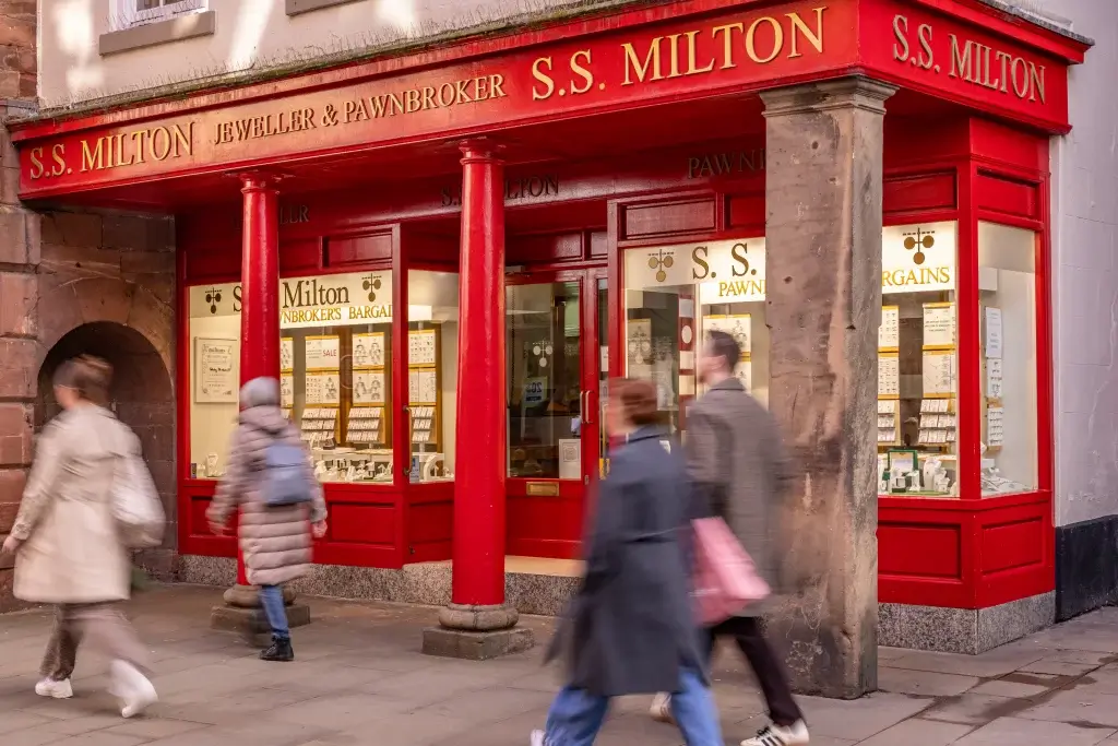 The bright red storefront of "S.S. Milton Jeweller & Pawnbroker," featuring display cases of jewelry, with two people walking by.