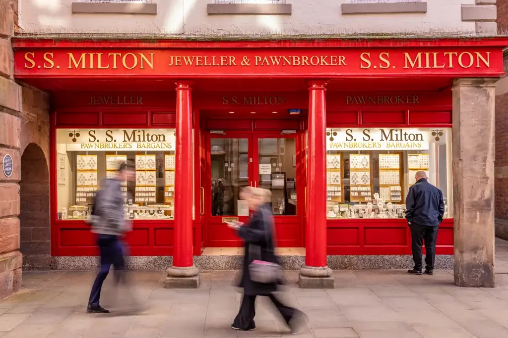 The bright red storefront of "S.S. Milton Jeweller & Pawnbroker," featuring display cases of jewelry, with two people walking by.