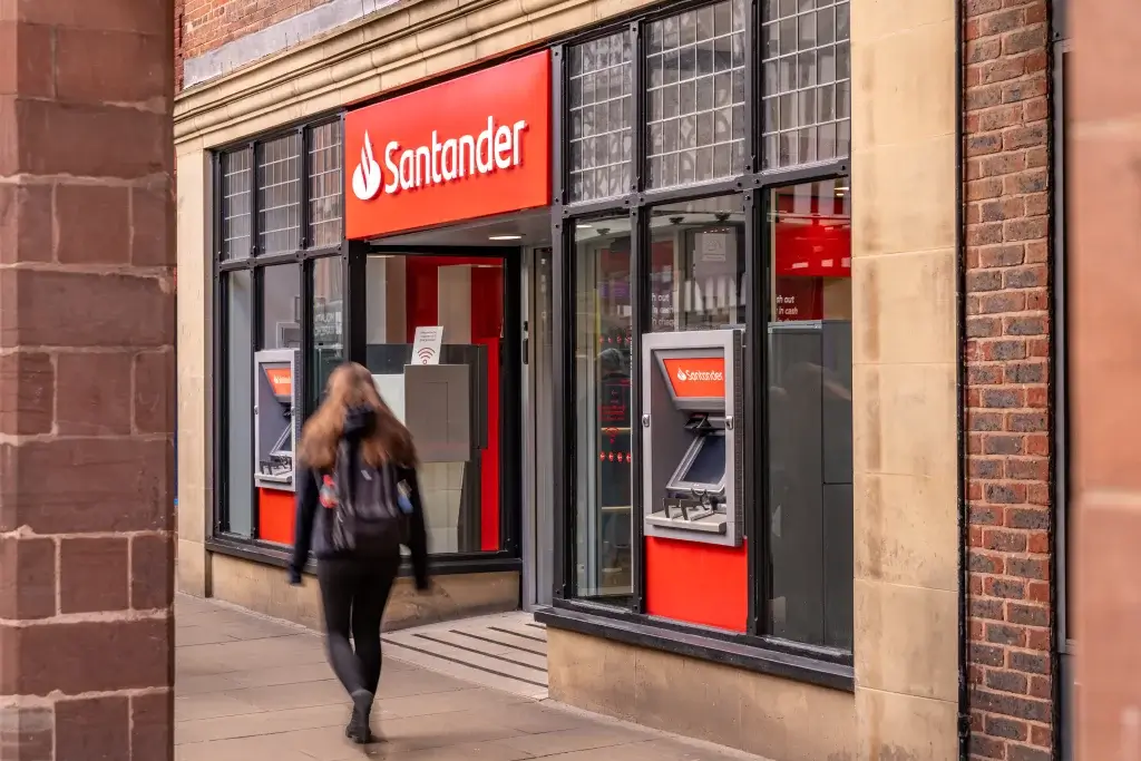 A person walks past a Santander bank with a bright red sign and ATM, set in a historic building.