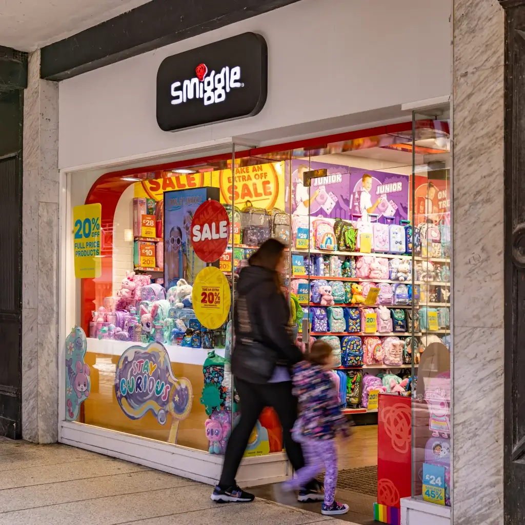 A woman and child walk past a vibrant Smiggle store, showcasing colorful bags, toys, and sale signage in a shopping area.