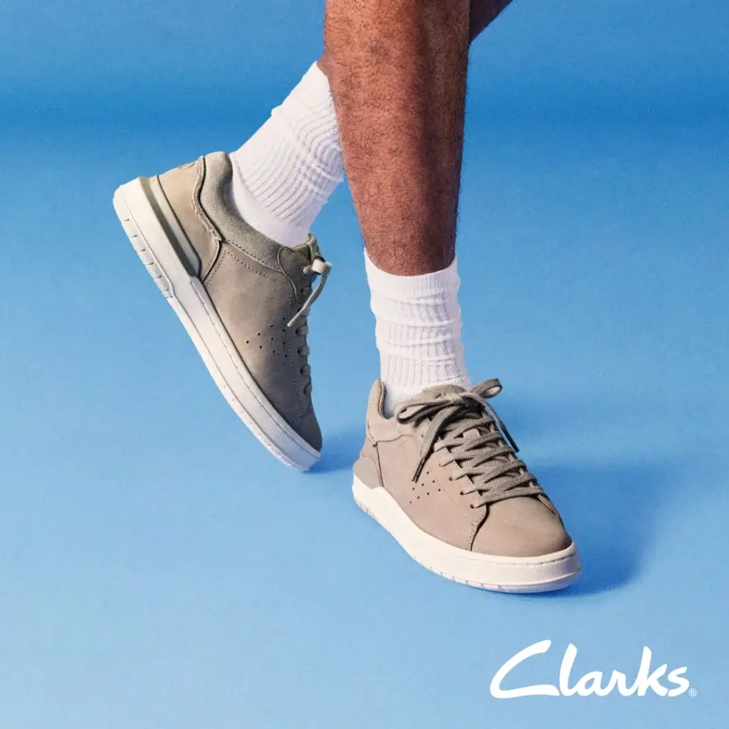 A close-up of a person wearing light gray Clarks sneakers and white socks, set against a soft blue backdrop.