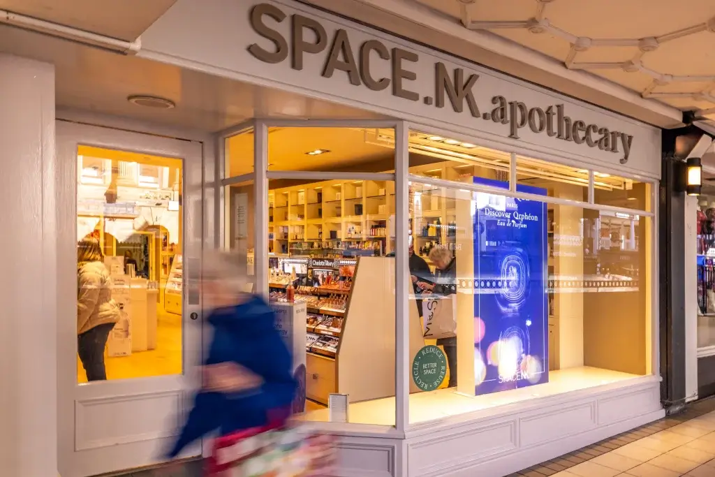 A blurred figure walks past the storefront of Space.NK Apothecary, showcasing beauty products and an inviting interior.