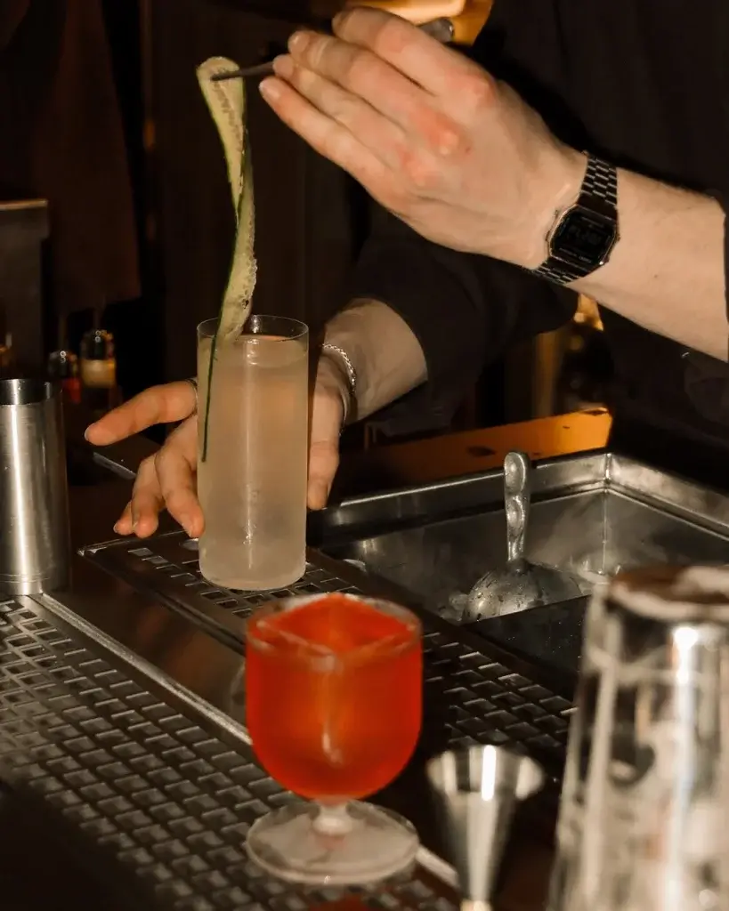 A bartender garnishes a clear cocktail with a cucumber slice while another vibrant red drink sits nearby on the bar.