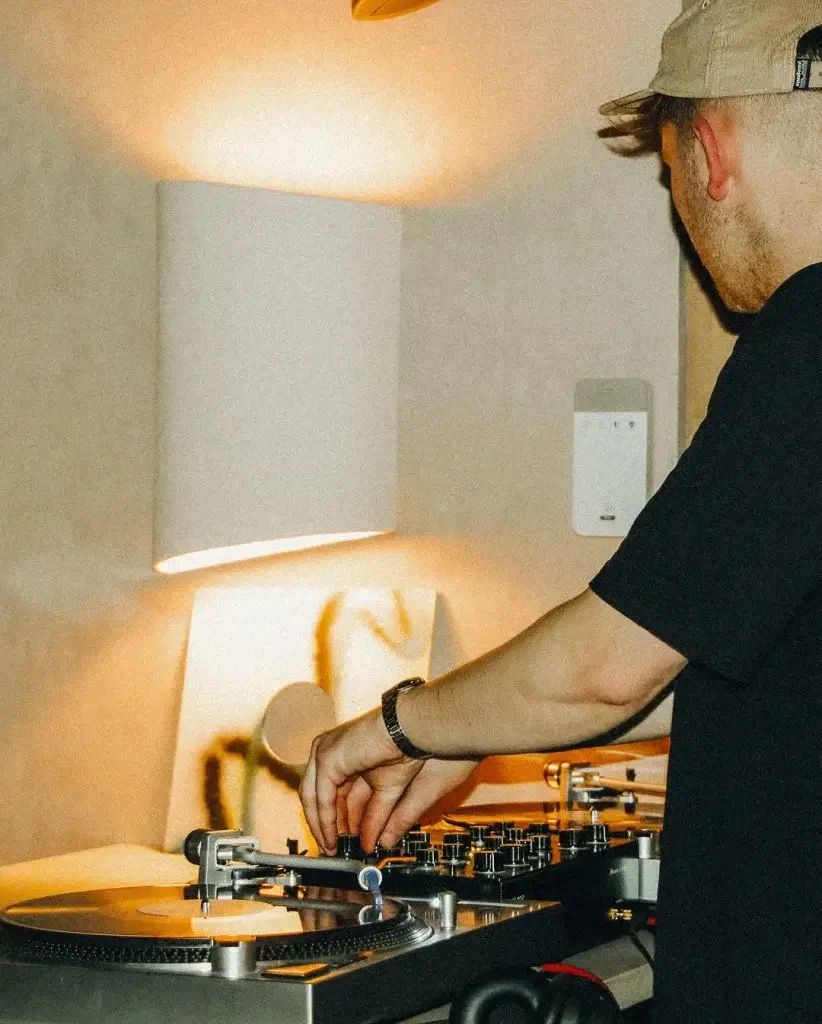 A DJ in a black t-shirt adjusts the turntable and mixer under soft ambient lighting, focused on creating music.