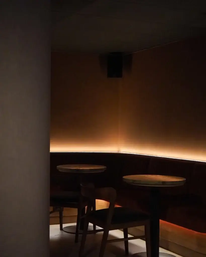 Intimate, dimly lit lounge with two round tables and plush seating, featuring soft, warm lighting along the walls.