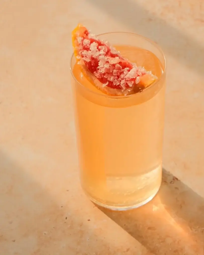 A refreshing cocktail in a clear glass, garnished with salt and a pink grapefruit slice, resting on a light, textured surface.