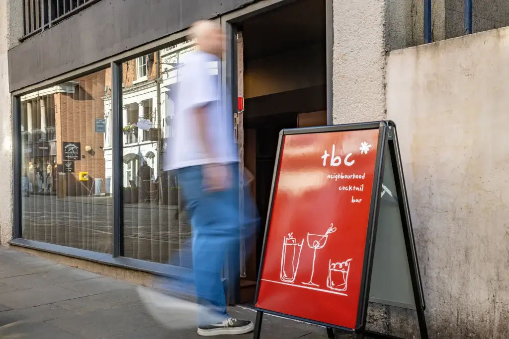 A blurred figure walks past a bar entrance labeled "tbc neighbourhood cocktail," with reflections of nearby buildings in the glass.