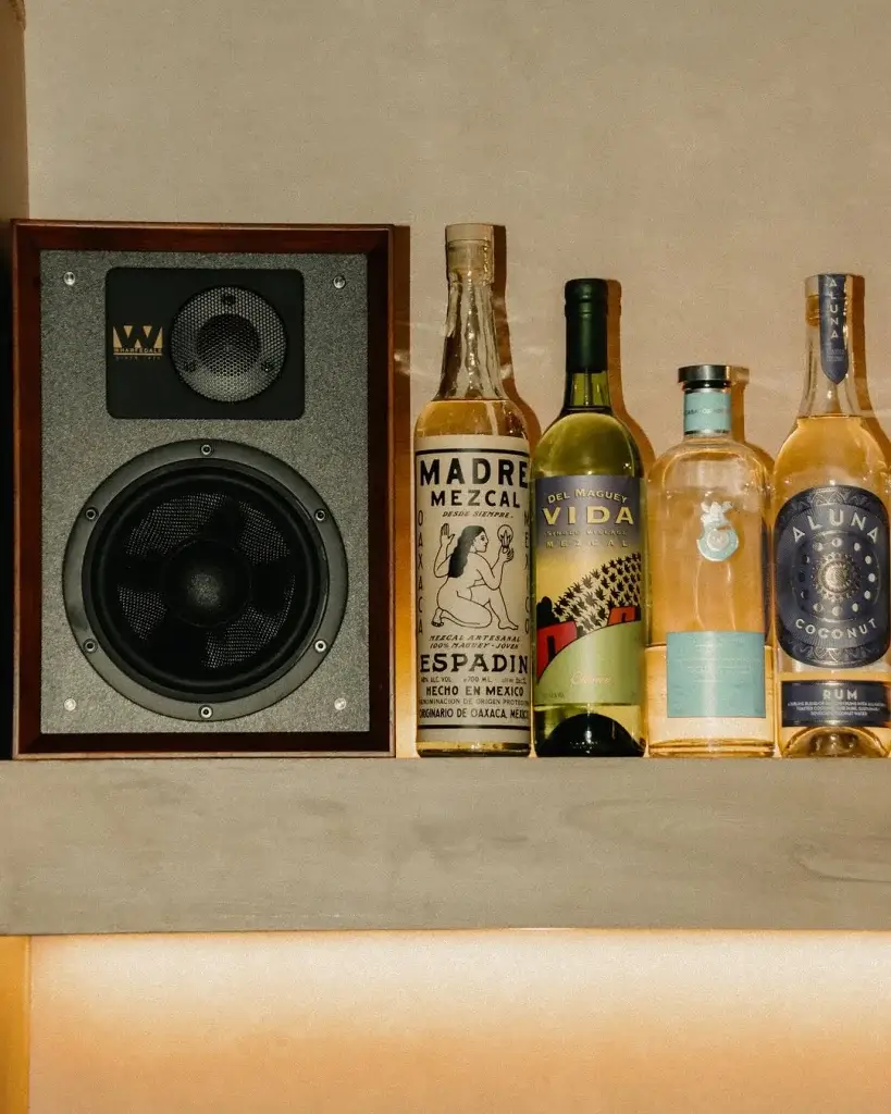 A wooden speaker next to four bottles of mezcal and rum on a shelf, illuminated by soft lighting from below.
