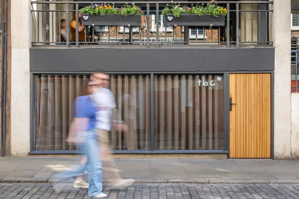 Two blurred pedestrians walk past a shop with a "tbc*" sign on the window, surrounded by flowers on the balcony above.