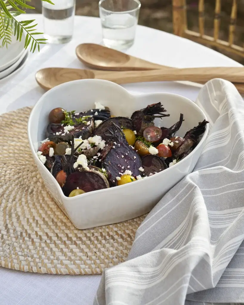 A heart-shaped bowl filled with roasted beets, cherry tomatoes, and feta cheese, set on a textured placemat with wooden utensils.