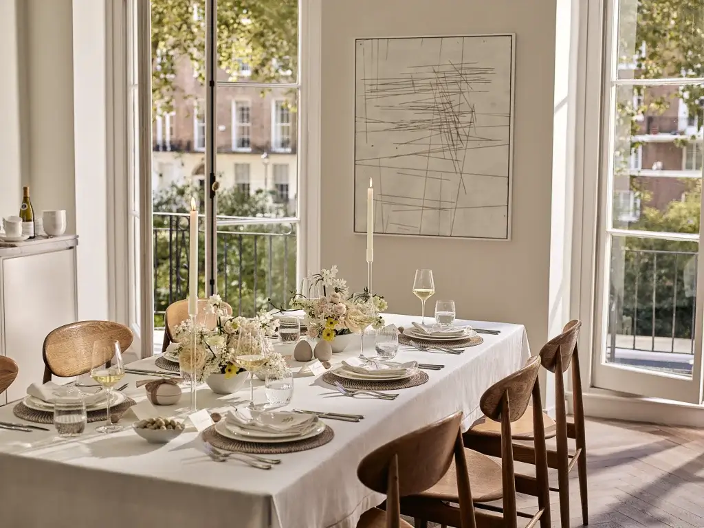 A beautifully set dining table with elegant tableware, flowers, and candles, overlooking a sunny outdoor view through large windows.