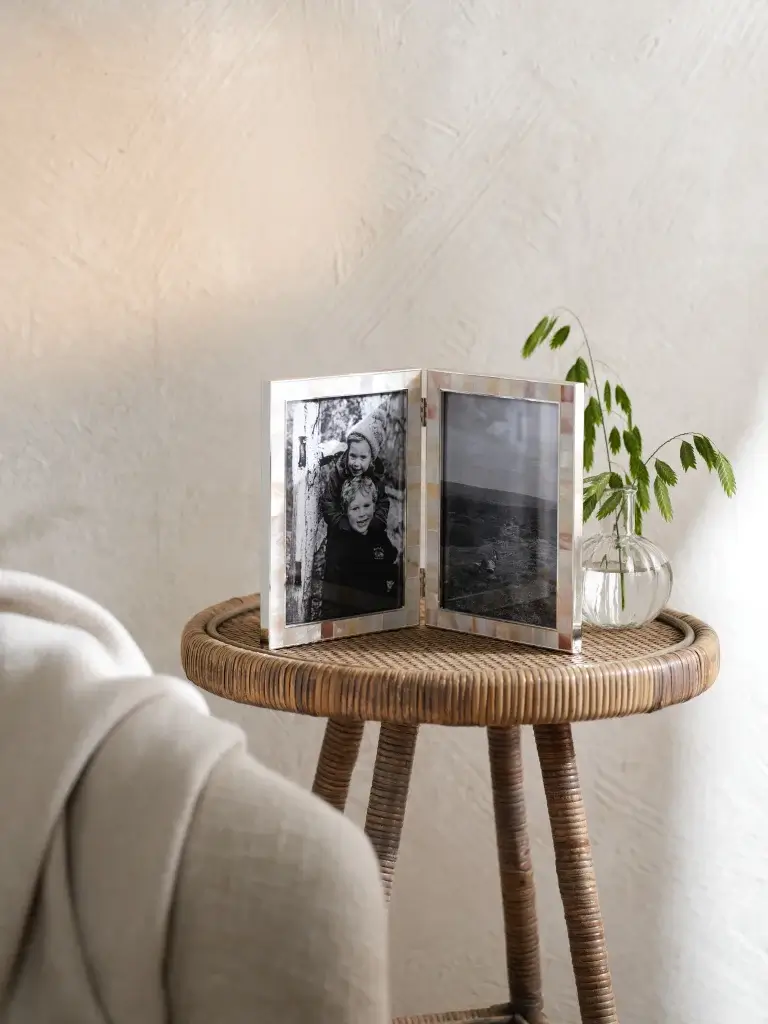 A decorative photo frame displaying two black-and-white images sits on a woven side table, alongside a small potted plant.