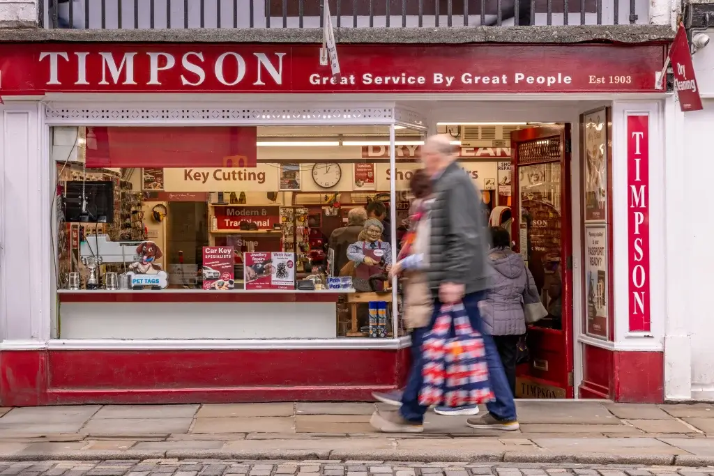 A busy Timpson store front with a red sign, showcasing services like key cutting, while customers walk outside.
