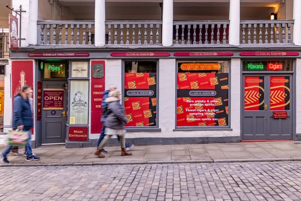 A tobacco shop with antique decor, featuring bold signage for cigars and spirits, as people walk by on a cobbled street.