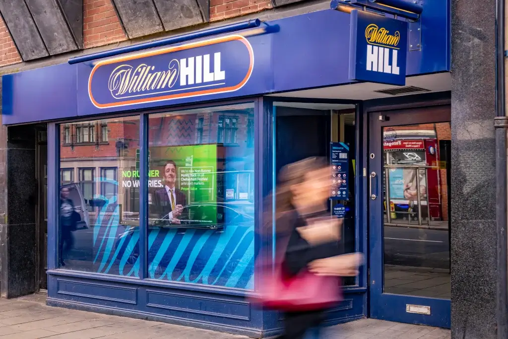 A William Hill betting shop displayed prominently, featuring digital advertisements and a customer passing by.