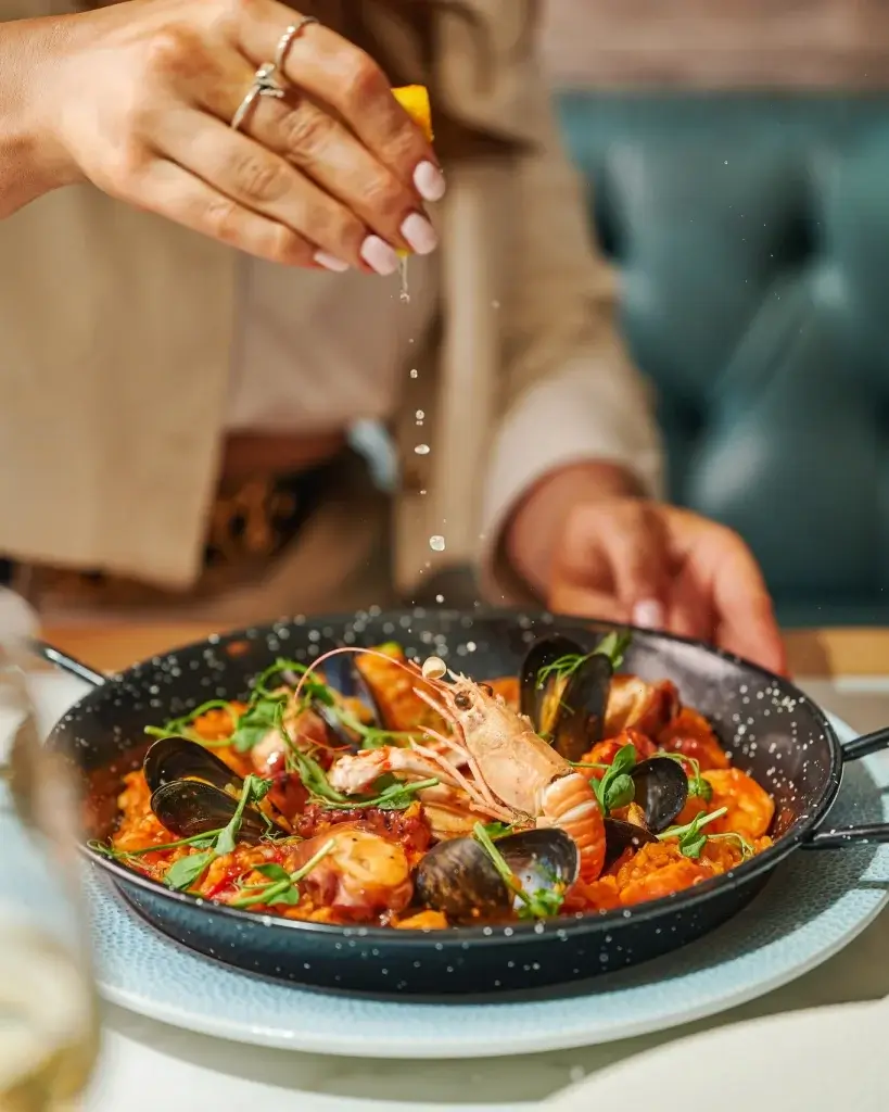 A hand squeezes lemon over a seafood paella, featuring shrimp, mussels, and vibrant herbs, in a black frying pan.