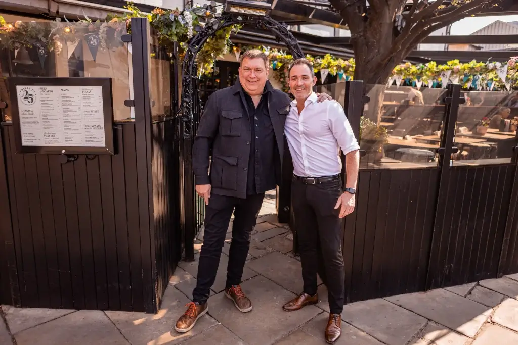 Two men stand together, smiling in front of a lush, decorated outdoor setting with greenery and festive bunting.