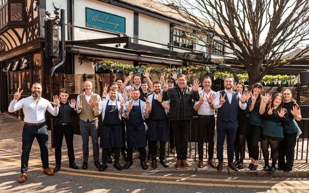 A group of smiling restaurant staff members is posing with cheerful hand gestures, celebrating an occasion outdoors against a festive backdrop.