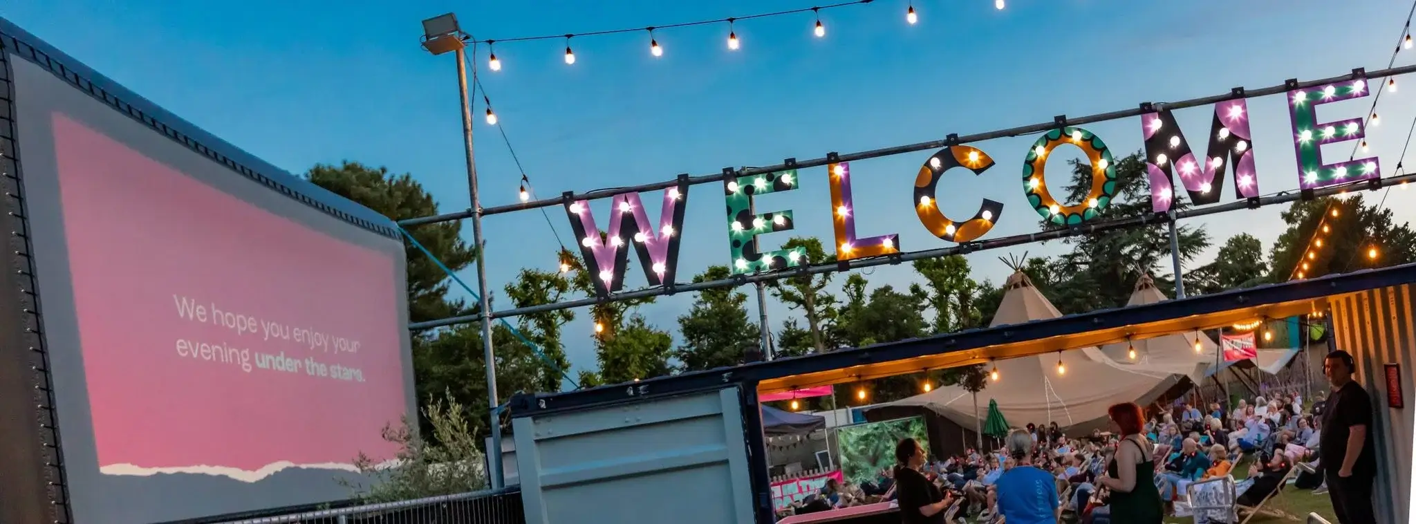 A vibrant "WELCOME" sign illuminated with colorful lights, set against a crowd enjoying an outdoor event under a twilight sky.