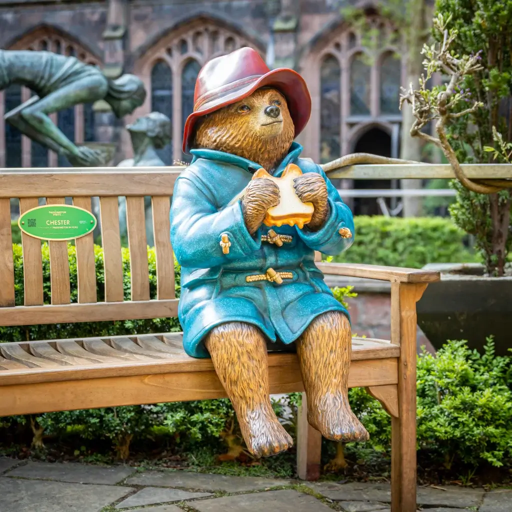 A life-sized statue of Paddington Bear in a blue coat and red hat sits on a bench, holding a sandwich, surrounded by greenery.
