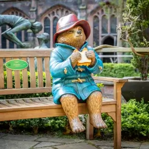 A life-sized statue of Paddington Bear in a blue coat and red hat sits on a bench, holding a sandwich, surrounded by greenery.