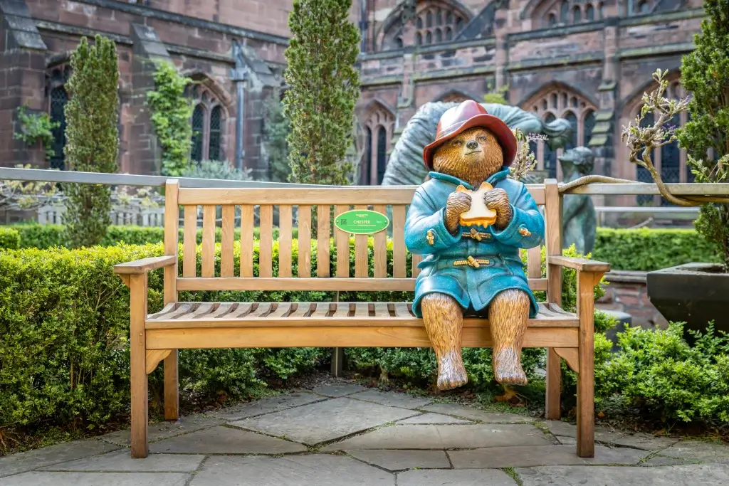 A life-sized statue of Paddington Bear in a blue coat and red hat sits on a bench, holding a sandwich, surrounded by greenery.
