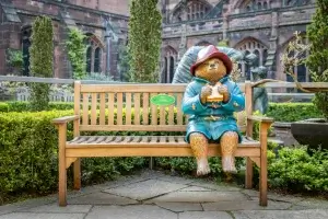A life-sized statue of Paddington Bear in a blue coat and red hat sits on a bench, holding a sandwich, surrounded by greenery.