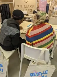 Two women sit at a table in a Primark store, engaged in a crafting activity, with bags promoting a sustainability initiative.